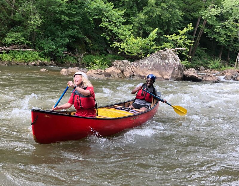 Two people are paddling a red canoe down a river. The person in the front is wearing a white helmet and a red life jacket, and is holding a blue paddle. The person in the back is wearing a black helmet and a red life jacket, and is holding a black paddle with a yellow blade. The river is surrounded by trees and rocks.